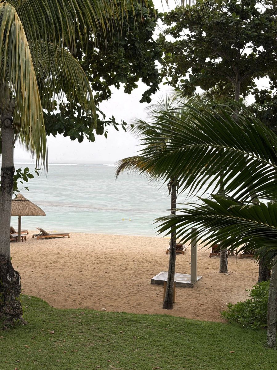 View from the St Regis gardens onto a near-empty beach with palm-thatched parasol and lagoon under a soft sky, Mauritius at Le Morne