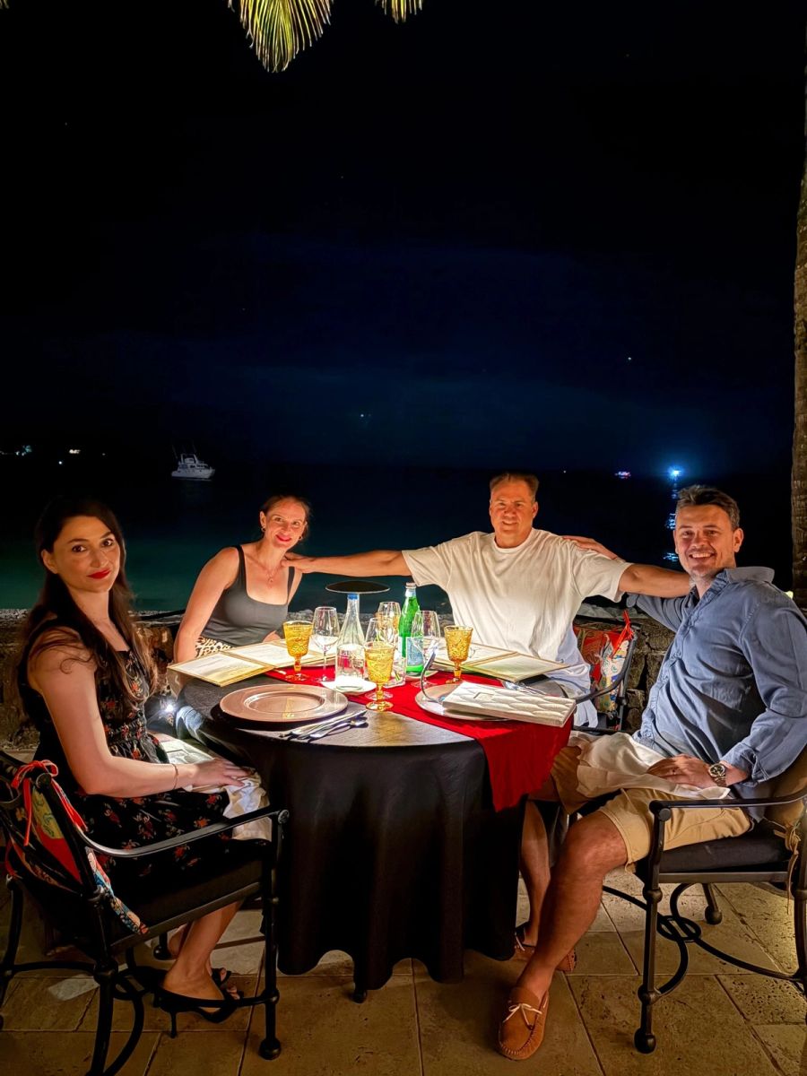 Two couples seated at an outdoor candlelit dinner table at Beachcomber Royal Palm, with the bay and boat lights visible in the background at night