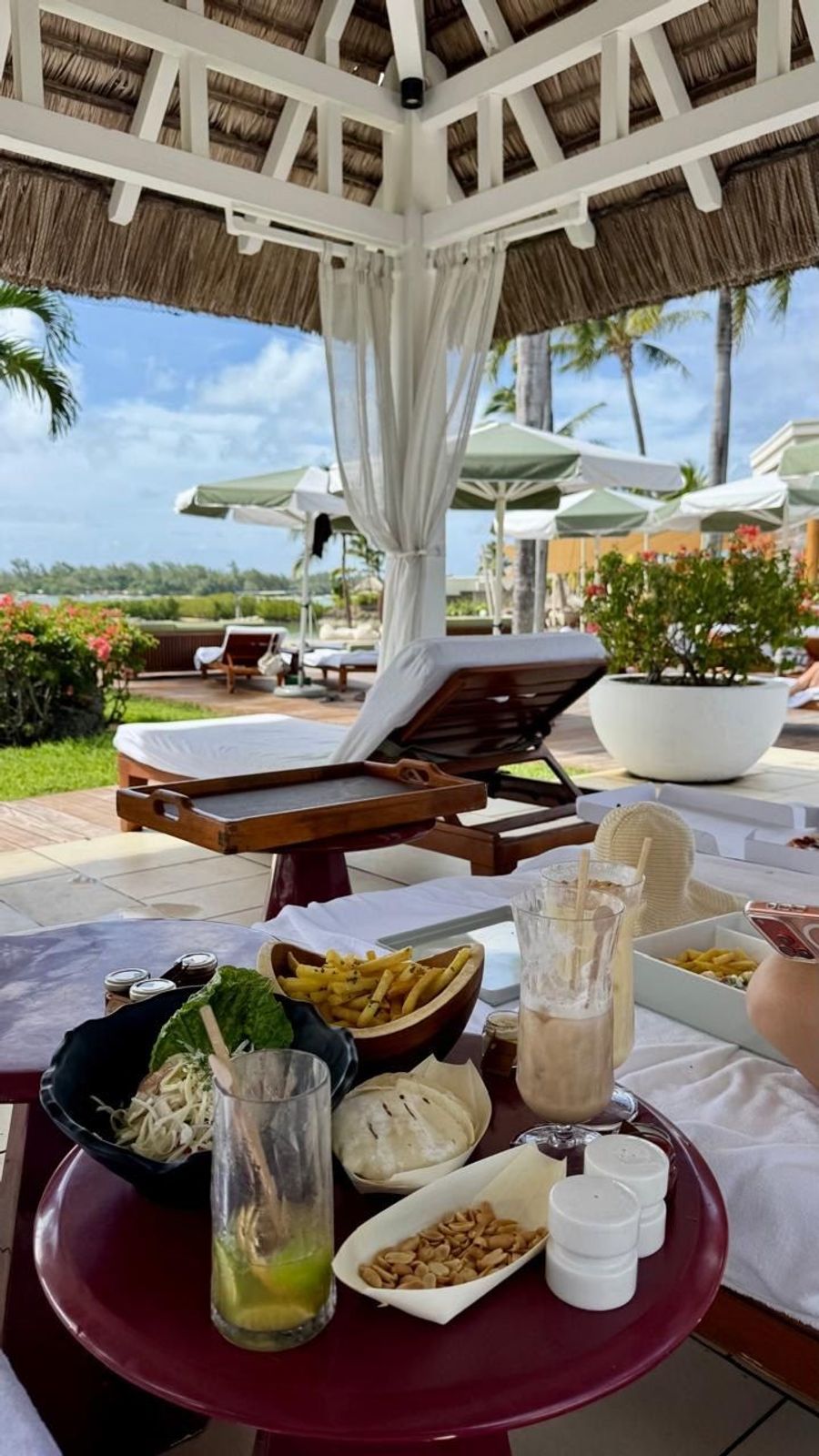 Cabana lunch table set with rice paper rolls, fries, mocktails and a tray on a daybed under a thatched roof, Four Seasons Mauritius at Anahita